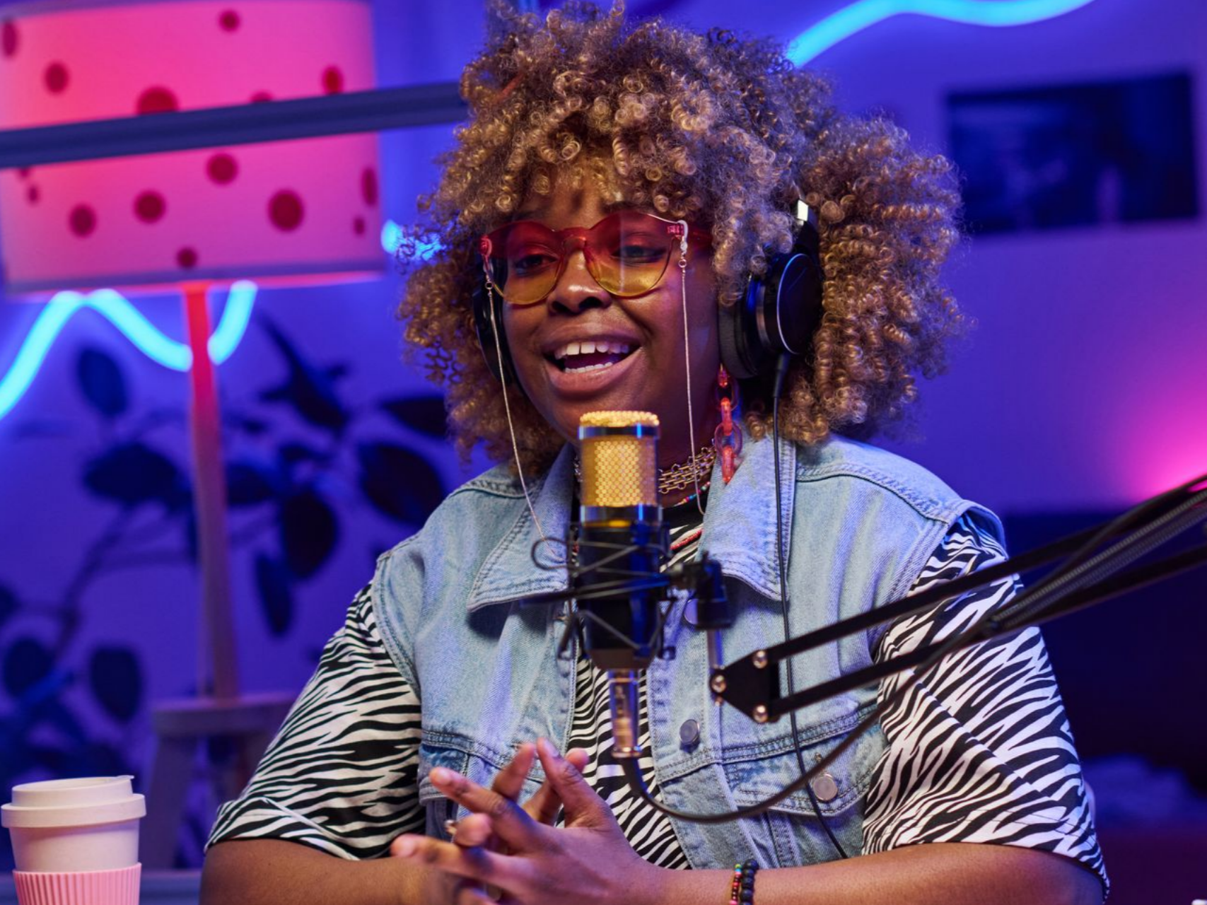 Une femme aux cheveux afro, portant des lunettes et un gilet en jean, parle dans un microphone. Des néons et une lampe à pois sont visibles en arrière-plan.