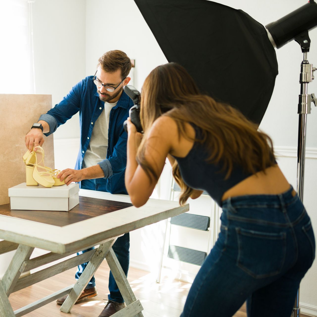 Deux photographes préparent une photo de talons jaunes. L'un dispose les chaussures, l'autre prend la photo, éclairée par une boîte à lumière.