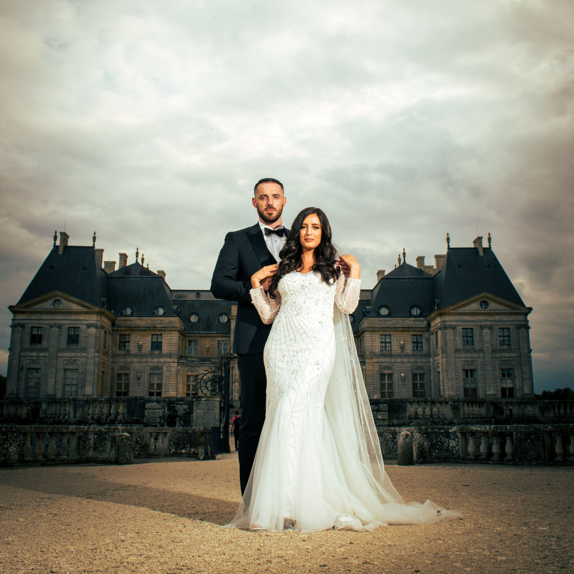 Les mariés posent devant un château. La mariée porte une robe blanche, le marié, un costume noir.