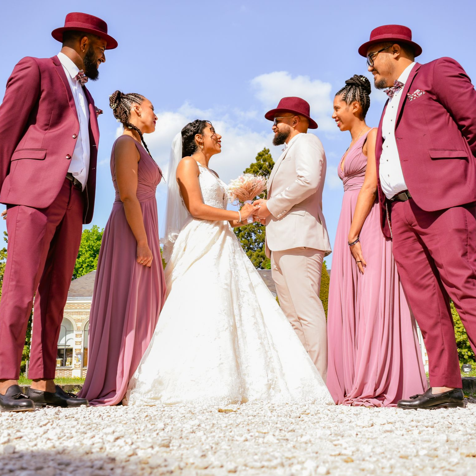 Cortège nuptial : les mariés se tiennent la main, entourés de demoiselles et garçons d’honneur vêtus de bordeaux et de rose, à l’extérieur.