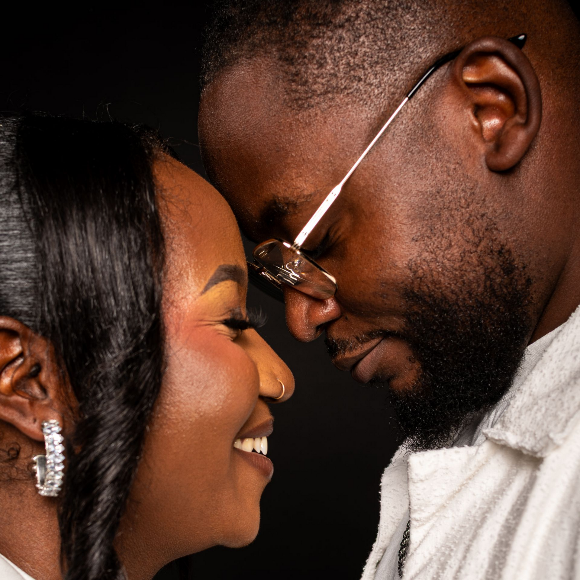 Couple souriant, front contre front, gros plan. Femme aux cheveux foncés et piercing au nez, homme barbu et portant des lunettes.