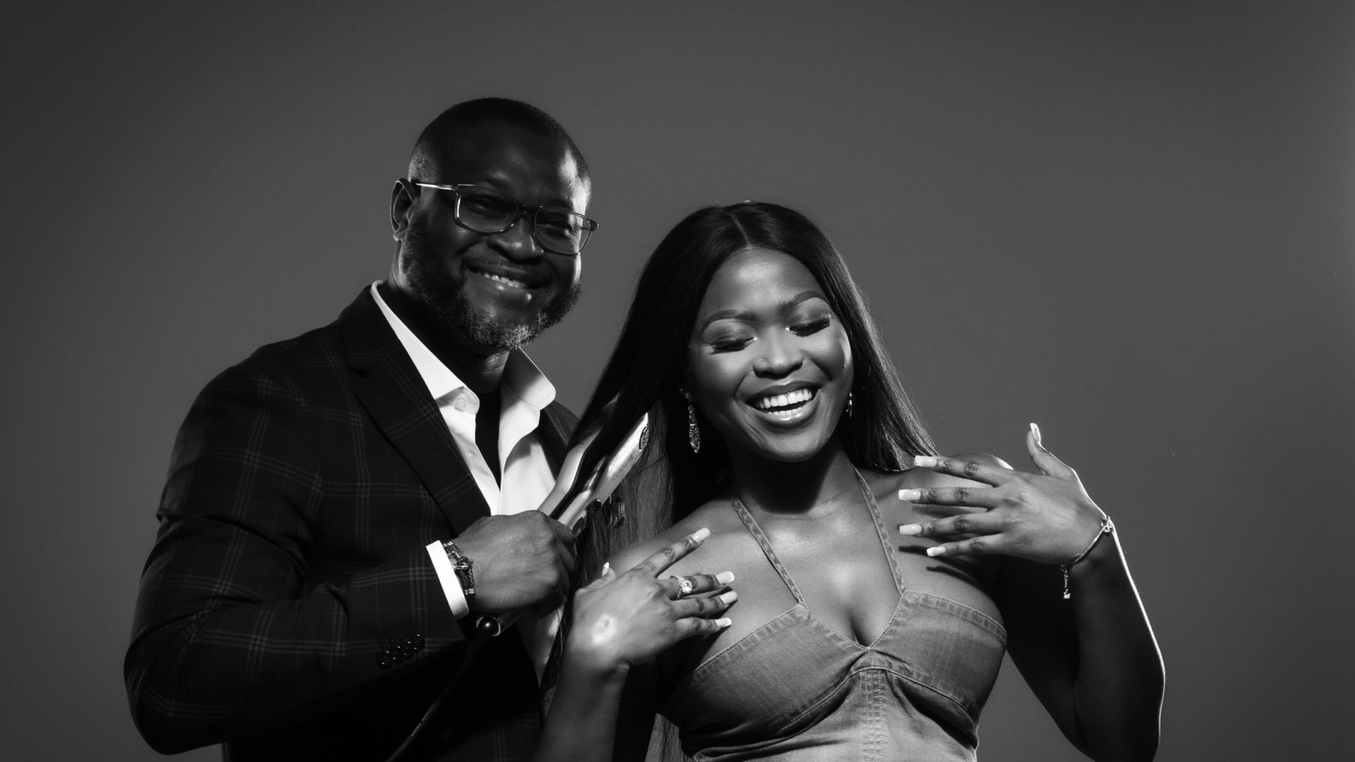 Un homme en costume et une femme en dos-nu sourient, posant côte à côte sur un fond uni. Noir et blanc.