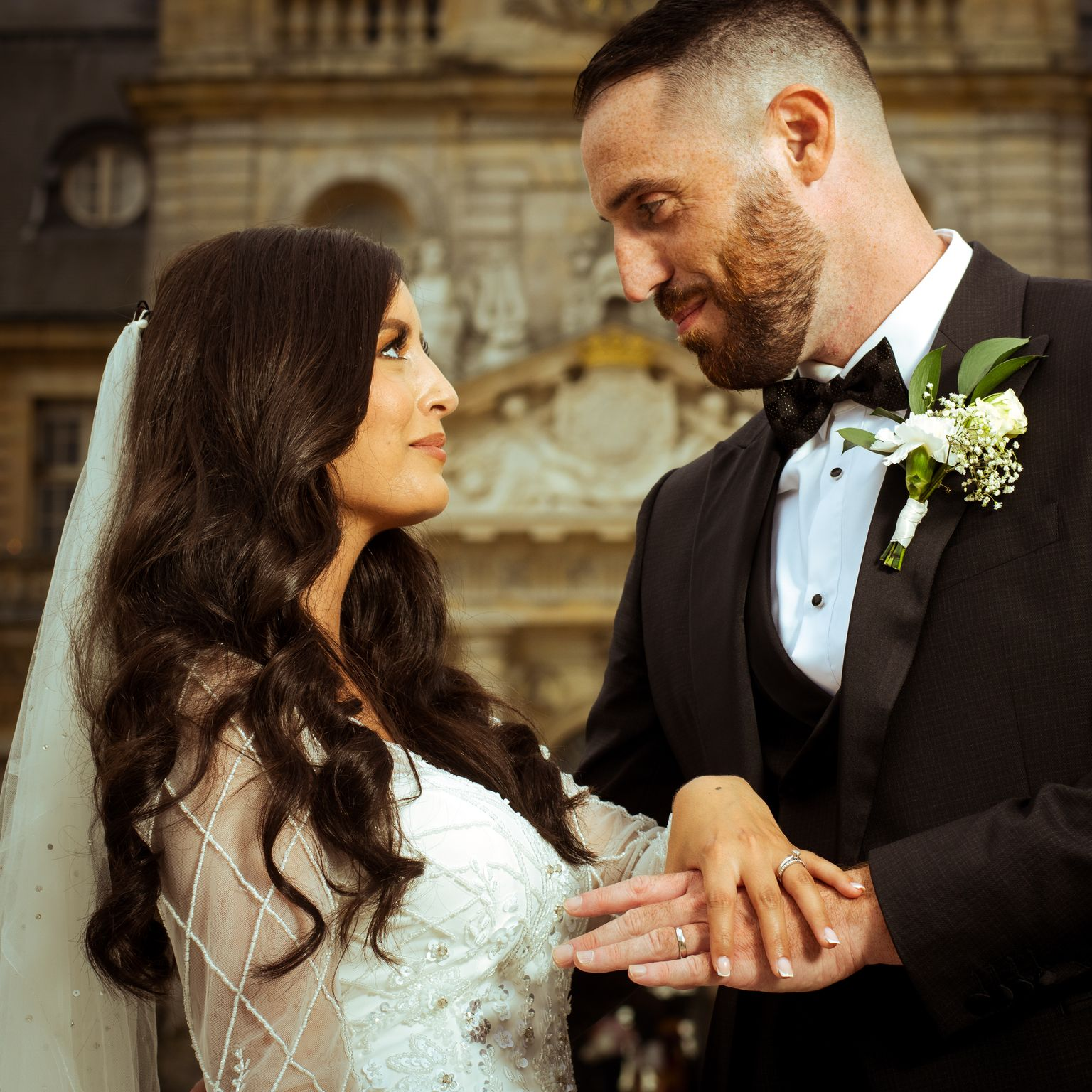 Les mariés se regardent. La mariée porte une robe blanche, le marié un smoking noir. Un bâtiment se dresse en arrière-plan.