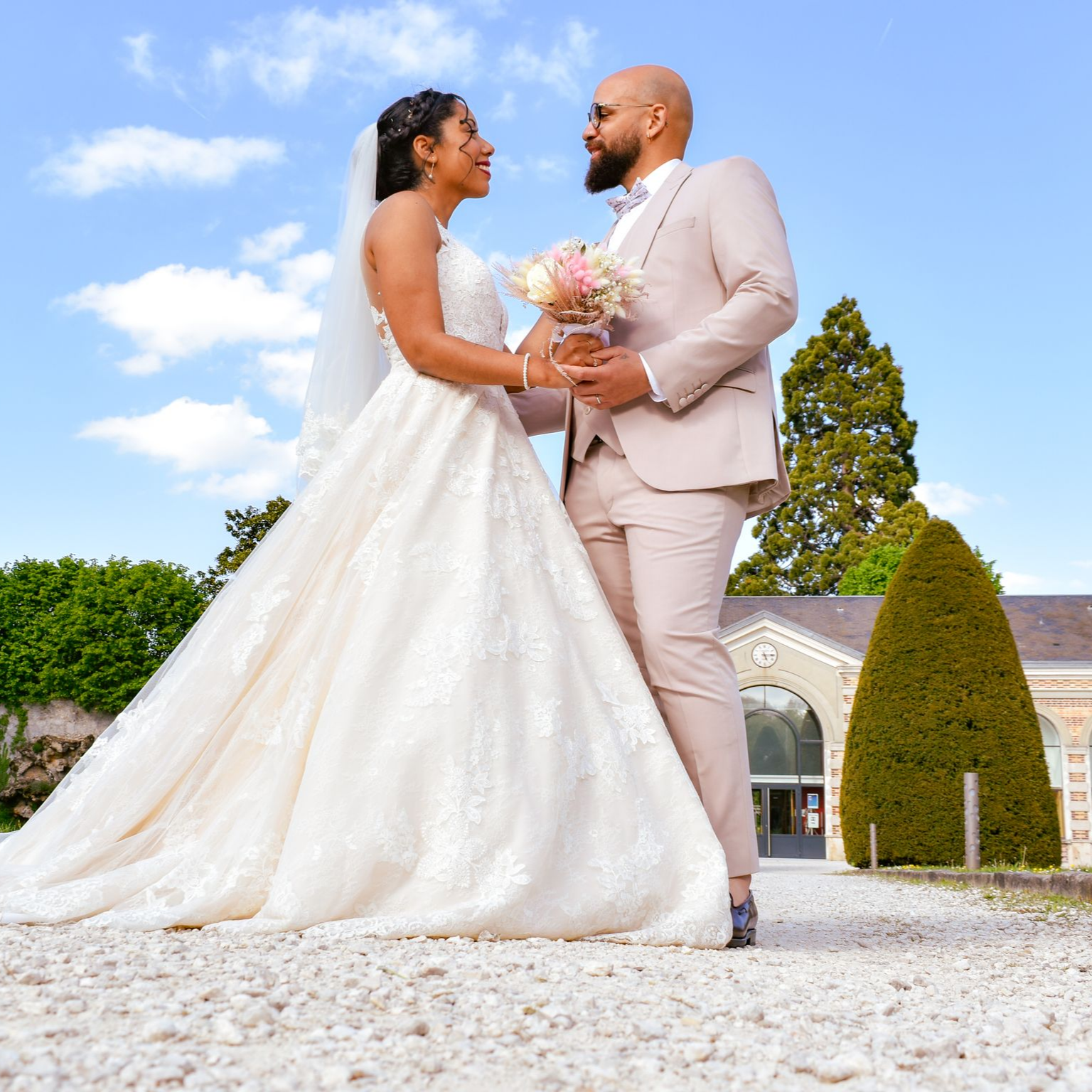 Un couple en tenue de mariage se regarde amoureusement. La mariée tient son bouquet. Le marié porte un costume clair. Vue extérieure avec un bâtiment.