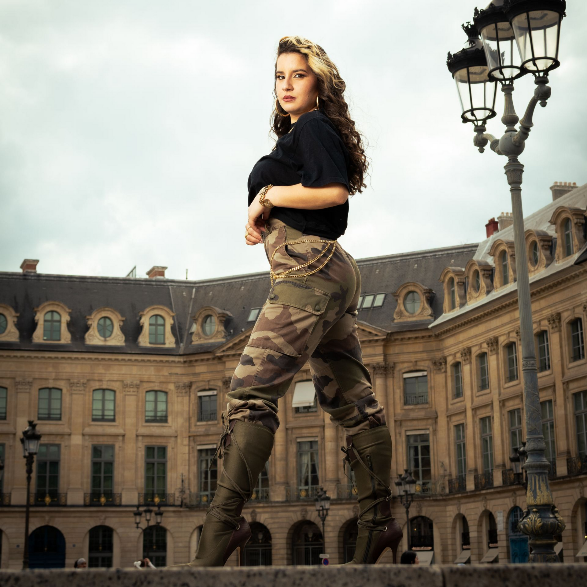 Une femme pose devant des bâtiments parisiens, vêtue d'un haut noir, d'un pantalon camouflage et de bottes.