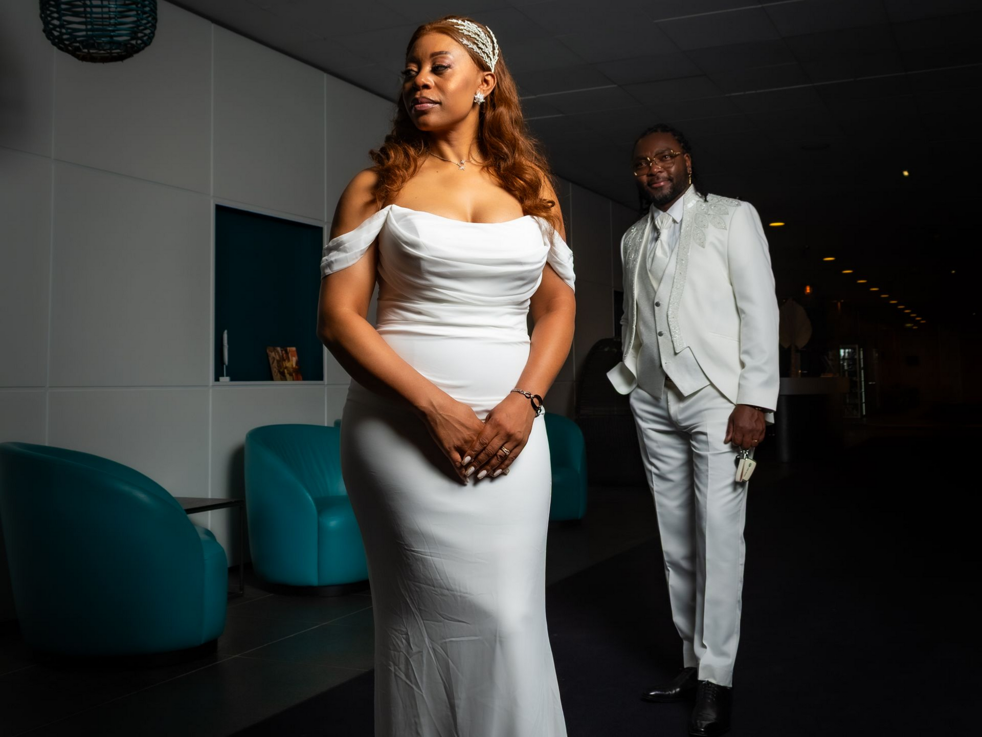 La mariée en robe blanche et le marié en costume blanc posent dans un intérieur moderne aux accents turquoise.