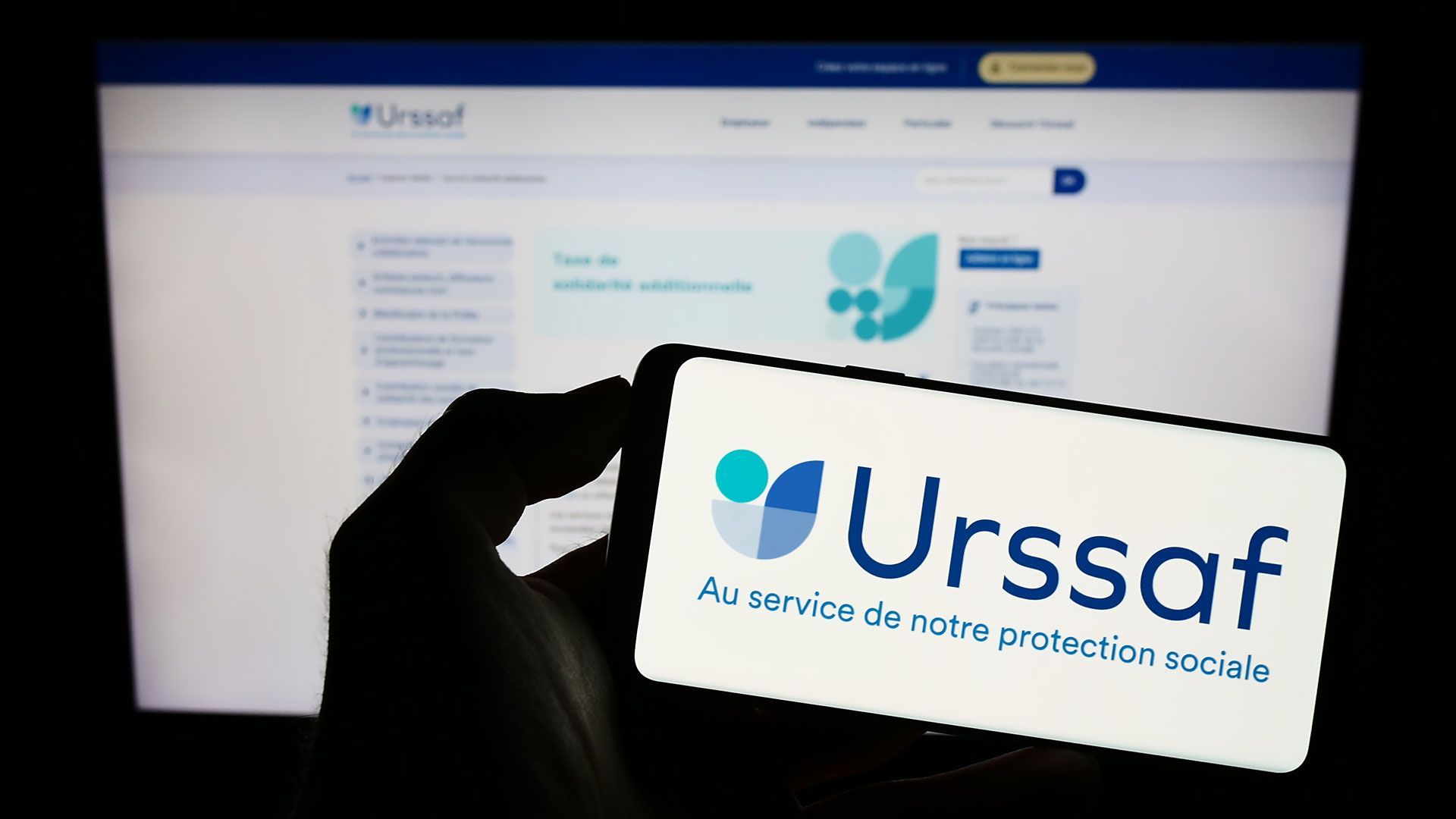 Une personne tenant un smartphone avec le logo Urssaf et le site en fond