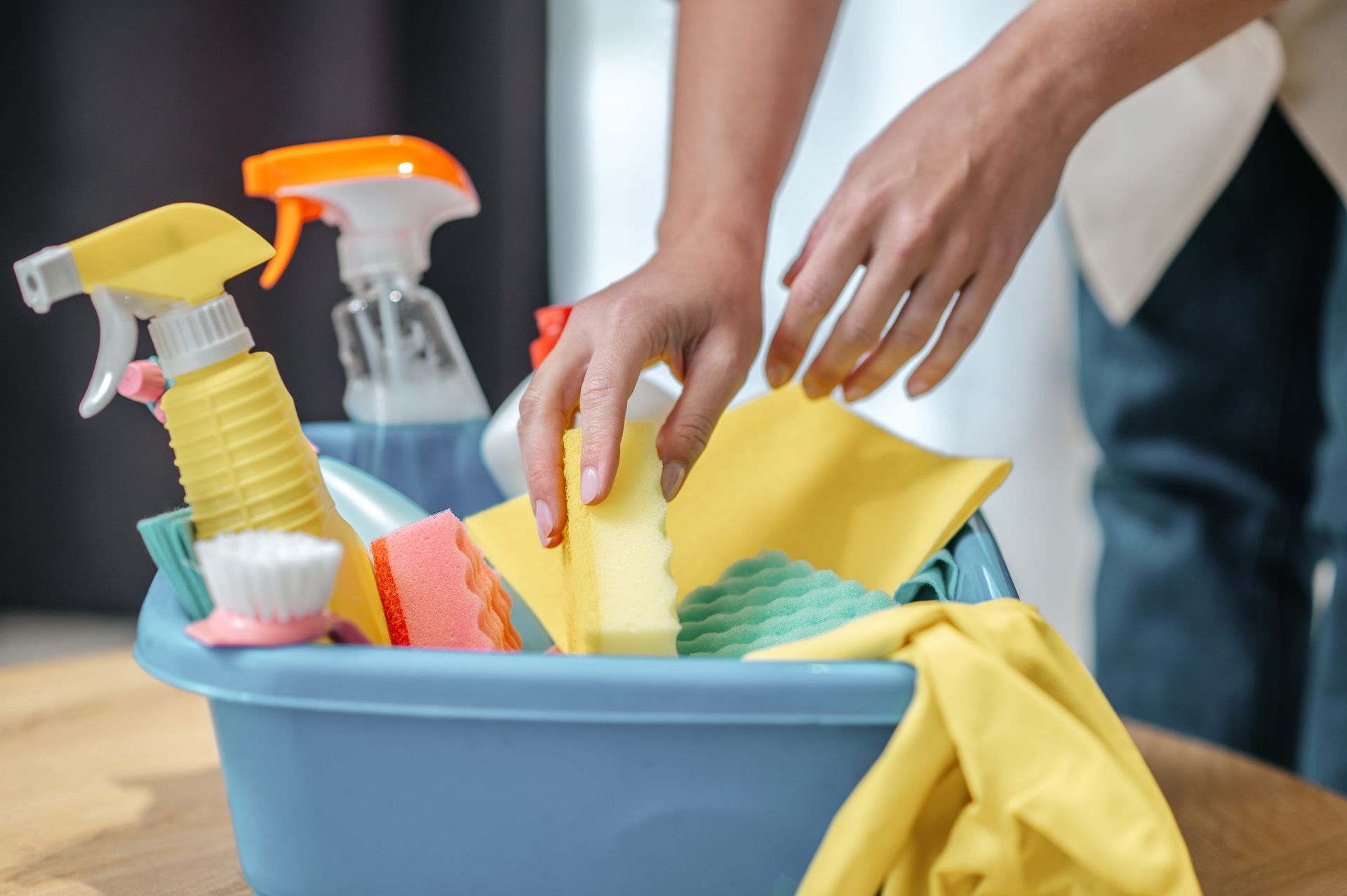 Manos limpiando un cubo azul con paños amarillos y botellas de spray cerca.