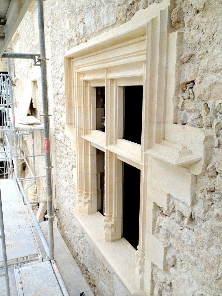 Construction d'un cadre de fenêtre en pierre sur un mur de pierre brute, échafaudages visibles.