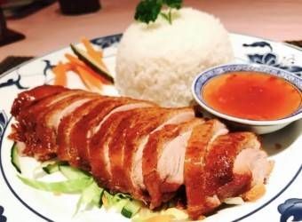 Gebratene Entenscheiben, serviert mit weissem Reis, Gurkenscheiben und einer kleinen Schüssel rötlicher Dip-Sauce auf einem gemusterten Teller.