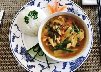 Eine Schüssel rotes Curry mit Gemüse neben einer Kugel weissem Reis auf einem Teller mit Blumenmuster.