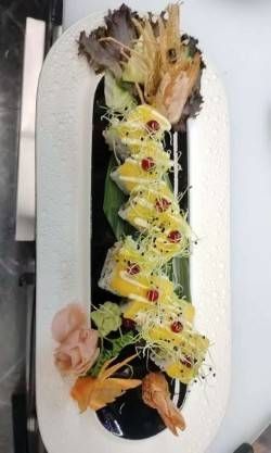 Eine Reihe Sushi-Rollen, garniert mit Mangoscheiben, einem Dressing und Microgreens, serviert auf einem länglichen weissen Teller mit Garnierung.