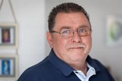 Ein Mann mit Brille und blauem Hemd steht vor einer Wand mit Bildern daran.