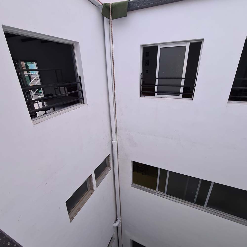 Mirando hacia arriba, a las paredes blancas del patio interior de un edificio. Hay ventanas, algunas con rejas negras. Una tubería blanca corre verticalmente.