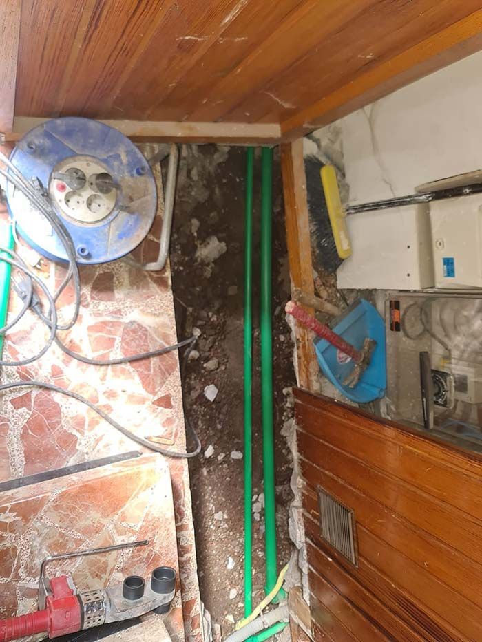 Una escena de construcción con tuberías verdes en una zanja. Hay herramientas y equipos eléctricos en una habitación con suelo de baldosas y paneles de madera.