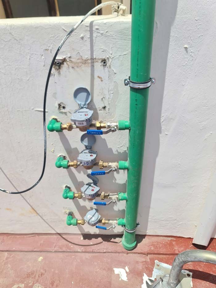 Contadores de agua y tuberías montados en una pared blanca, probablemente en el exterior. Se ven tuberías verdes, válvulas azules y contadores grises.