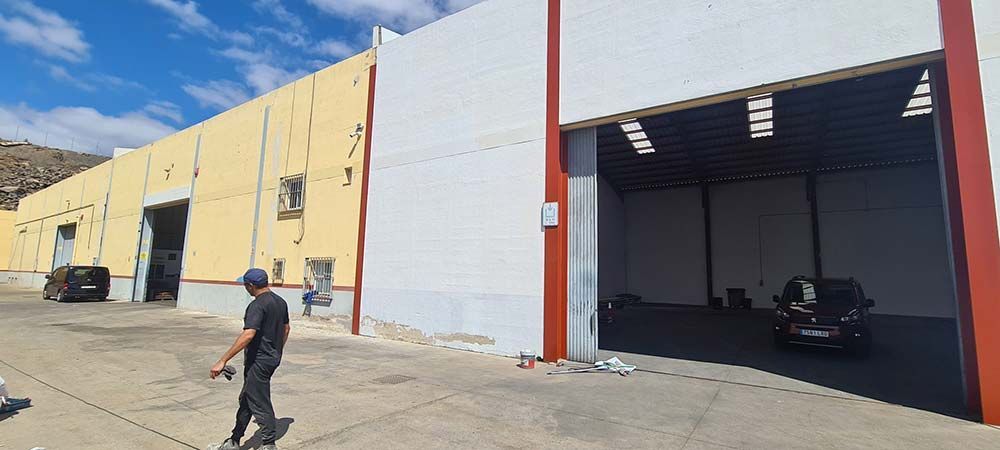 Un hombre pasa junto a un almacén con la puerta abierta y un coche oscuro aparcado dentro. El edificio tiene una fachada amarilla y blanca bajo un cielo azul.