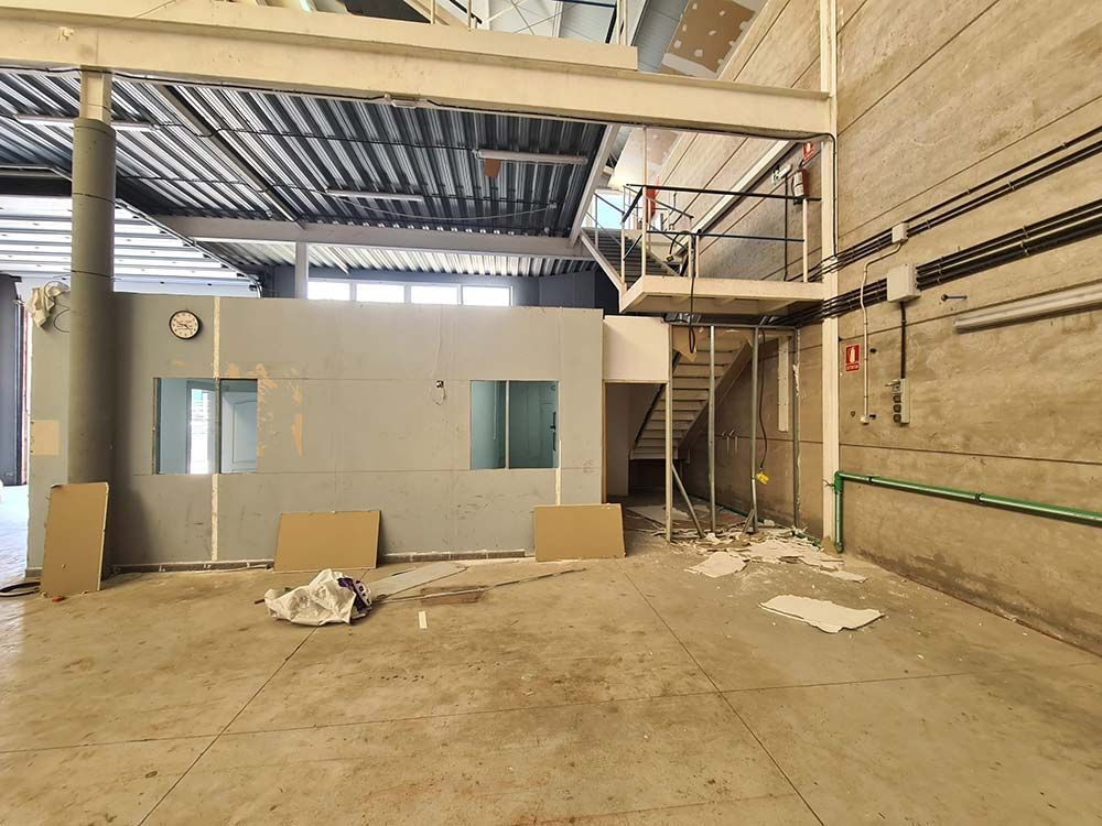 Vista interior de un edificio en construcción con vigas expuestas, una pared gris con ventanas y una escalera.