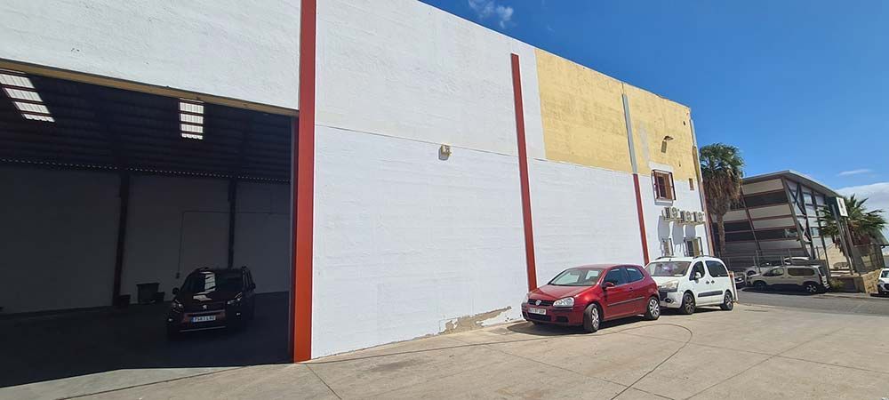Vista exterior de un almacén con coches aparcados. El edificio tiene paredes blancas y amarillas con molduras rojas y una gran entrada abierta.