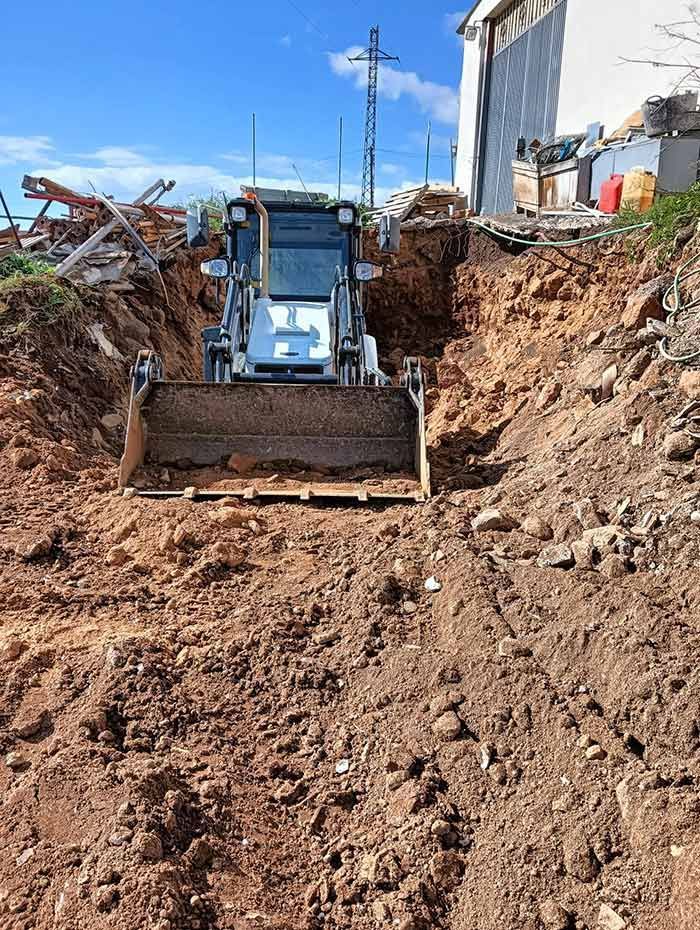 Una pequeña cargadora frontal en una zanja profunda y estrecha. La máquina, blanca y gris, excava en tierra marrón bajo un cielo azul.