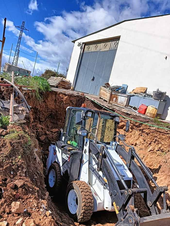 Minicargadora Bobcat blanca en una zanja, trabajando cerca de un edificio con una puerta azul. El cielo está nublado.