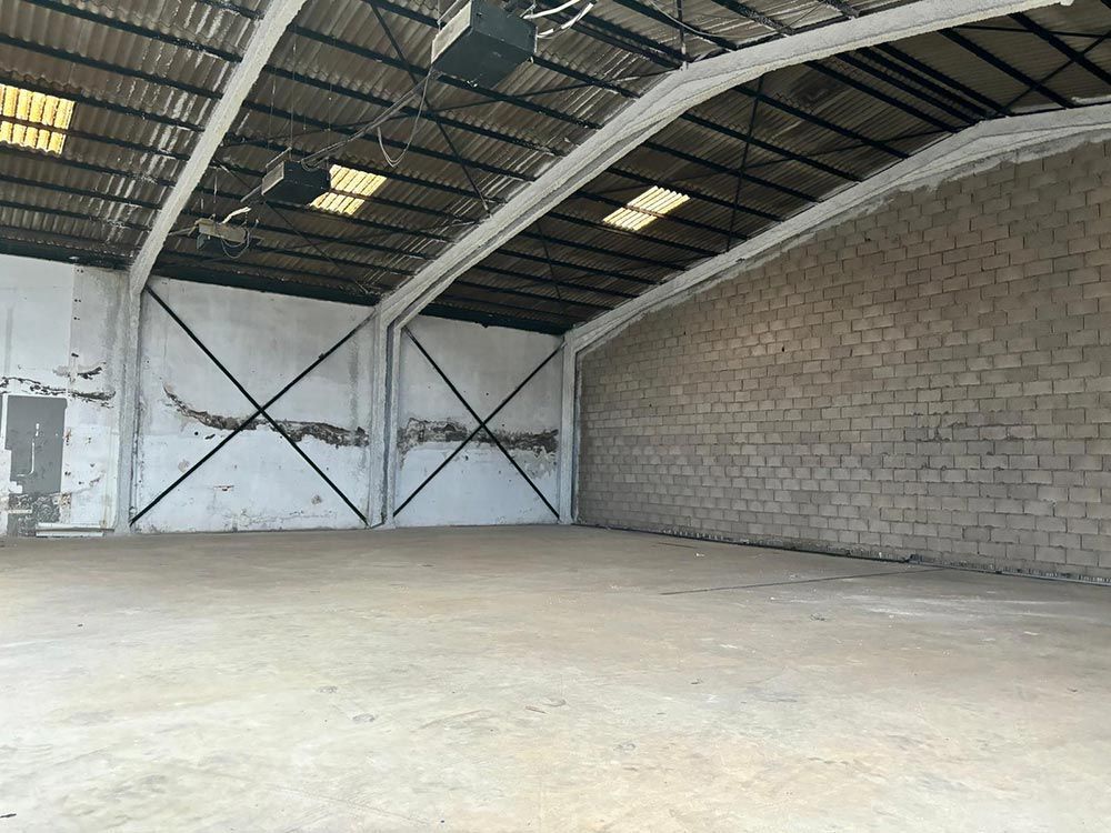 Interior de almacén vacío, piso de concreto, paredes de ladrillo y pintadas, techo de metal arqueado y tragaluces.