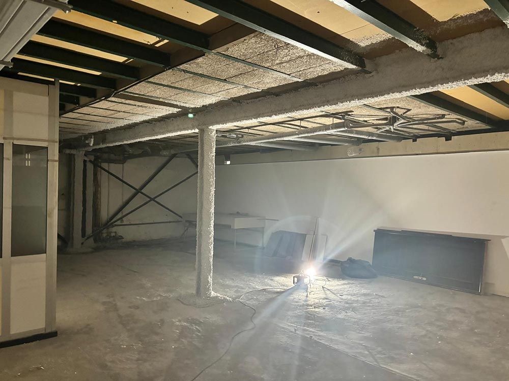 Un espacio interior inacabado con vigas metálicas, soportes y techo visto. Un foco ilumina el suelo de hormigón y las paredes blancas.