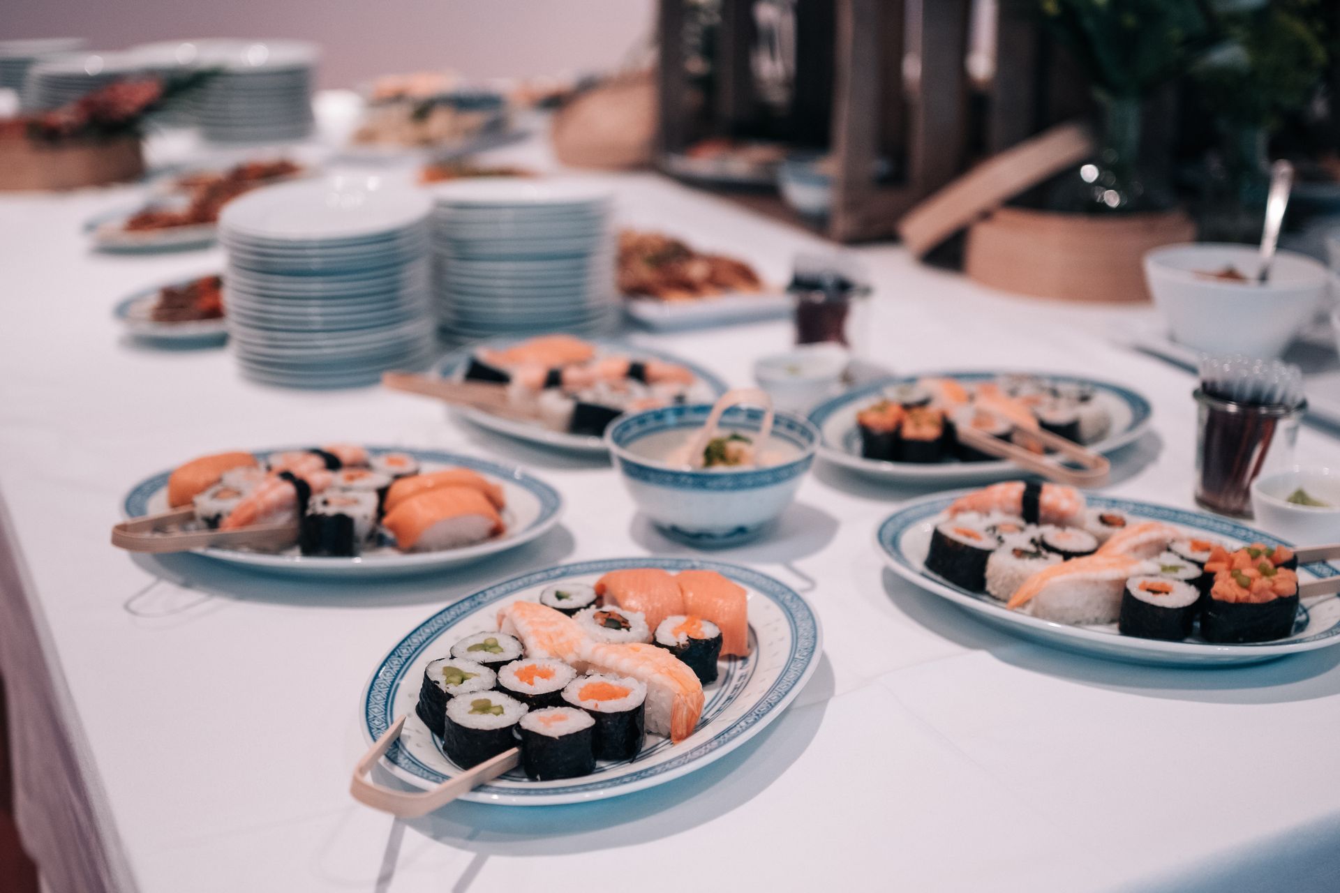 Sushi ist auf einem weißen Tisch ausgebreitet. Zu sehen sind Teller mit Sushi, Schüsseln und Tellerstapel.