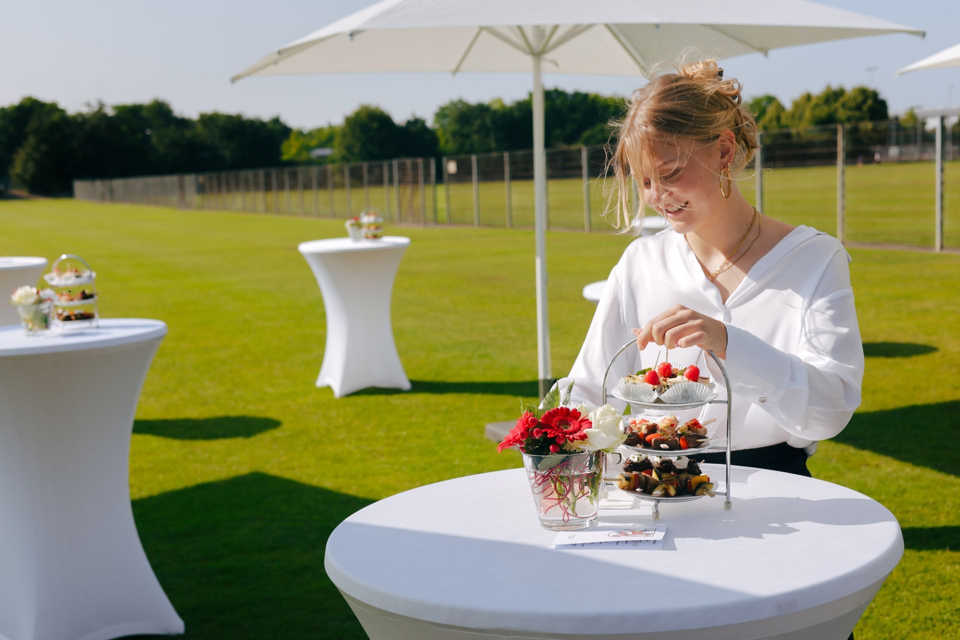 Frau arrangiert Gebäck auf einem Tisch im Freien. Weiße Sonnenschirme und Tische auf einer Wiese.