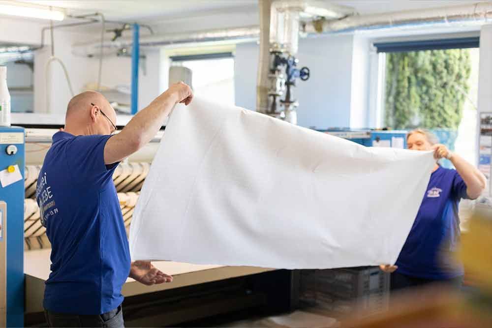 Zwei Männer halten in einem Waschsalon ein großes weißes Handtuch.