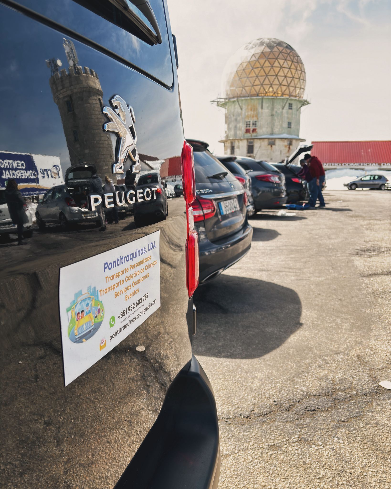 Furgão Peugeot preto com logotipo e adesivo, refletindo um prédio com cúpula dourada e carros estacionados.