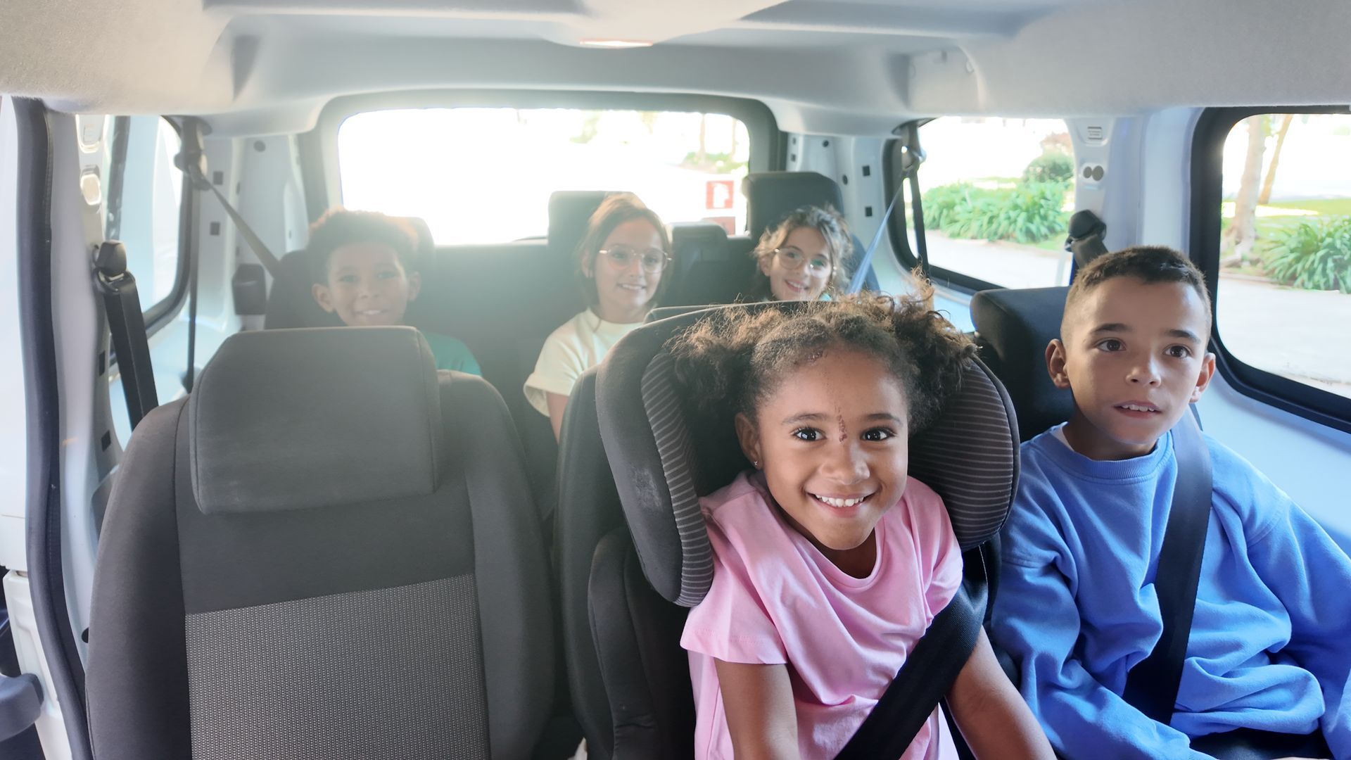 Cinco crianças sorrindo no banco de trás de uma van, algumas em cadeirinhas, todas usando cinto de segurança.