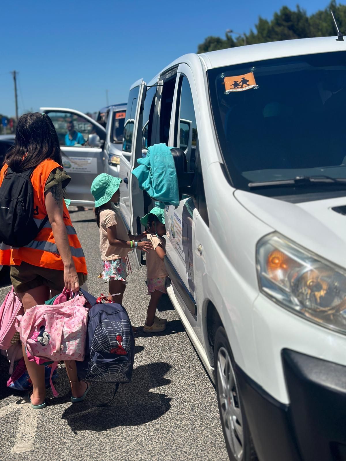 Uma mulher ajuda crianças a embarcarem em uma van branca. Uma das crianças usa um chapéu verde. Dia ensolarado e radiante.