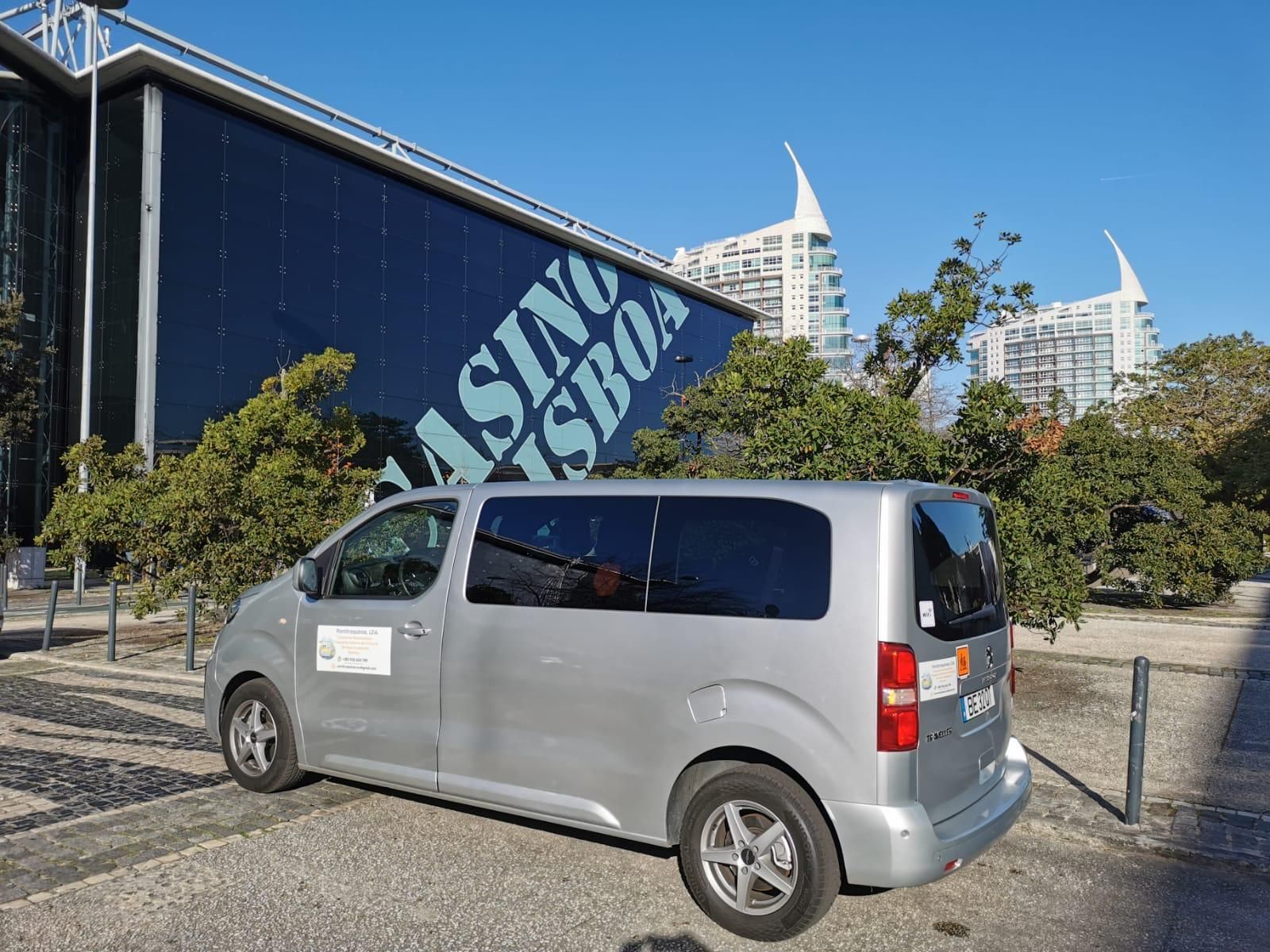 Uma carrinha prateada estacionada em frente ao Casino Lisboa, com arquitetura moderna e vista para a água.