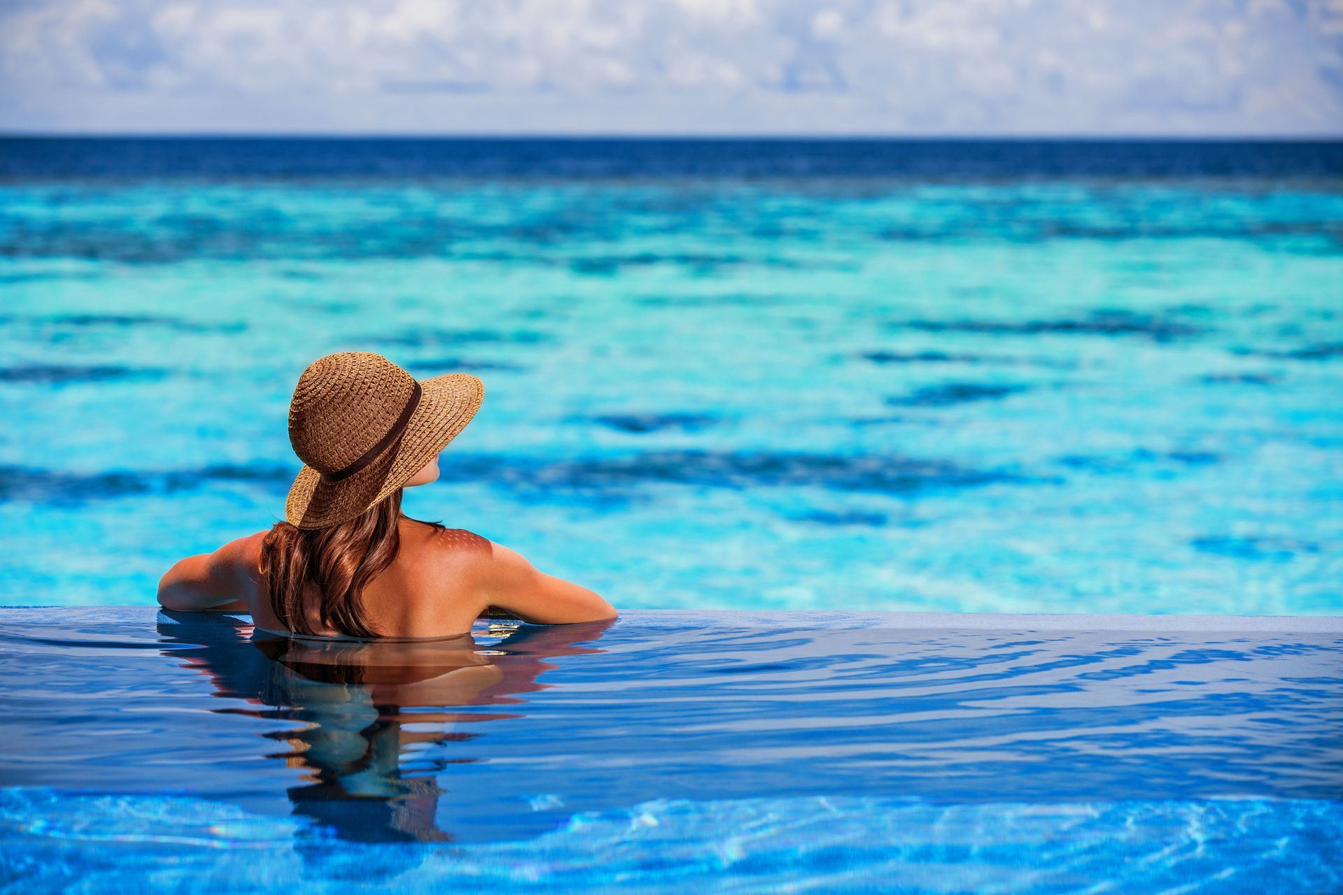 Person in a straw hat relaxing in a turquoise infinity pool facing the ocean
