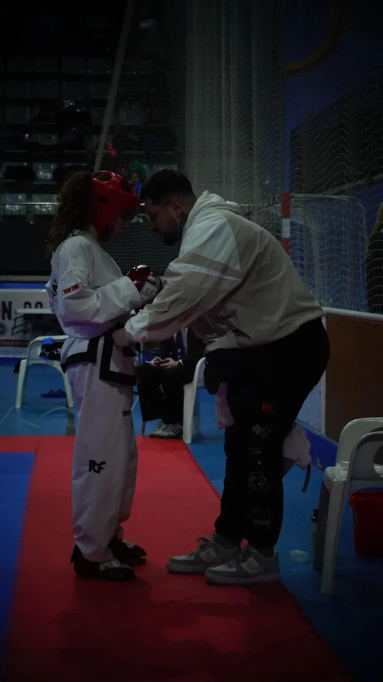 Un entrenador ajusta el uniforme de taekwondo de un alumno que lleva un casco rojo en una pista de competición.