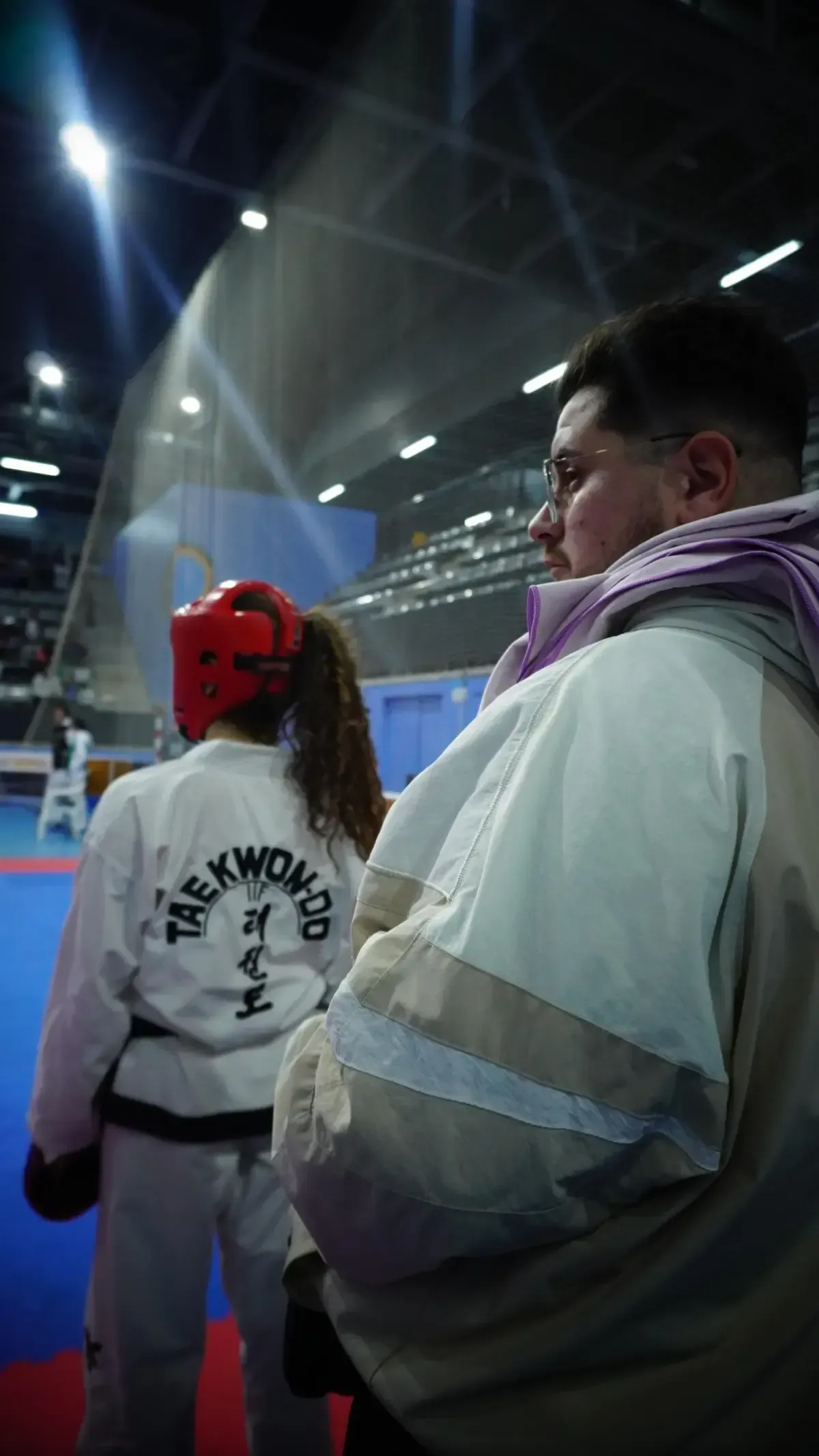 Una persona con uniforme de taekwondo y gorro rojo permanece de pie sobre una colchoneta azul