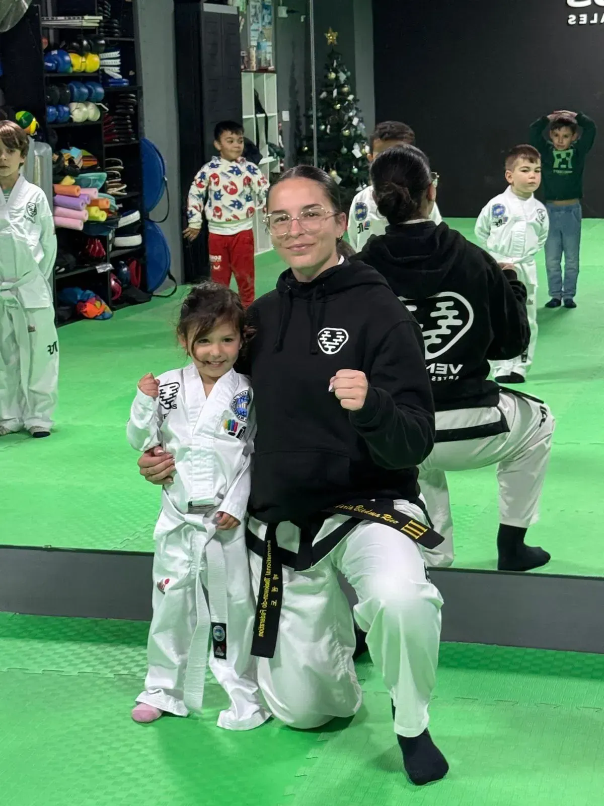 Un instructor de artes marciales con una sudadera negra con capucha y uniforme blanco está arrodillado junto a un estudiante