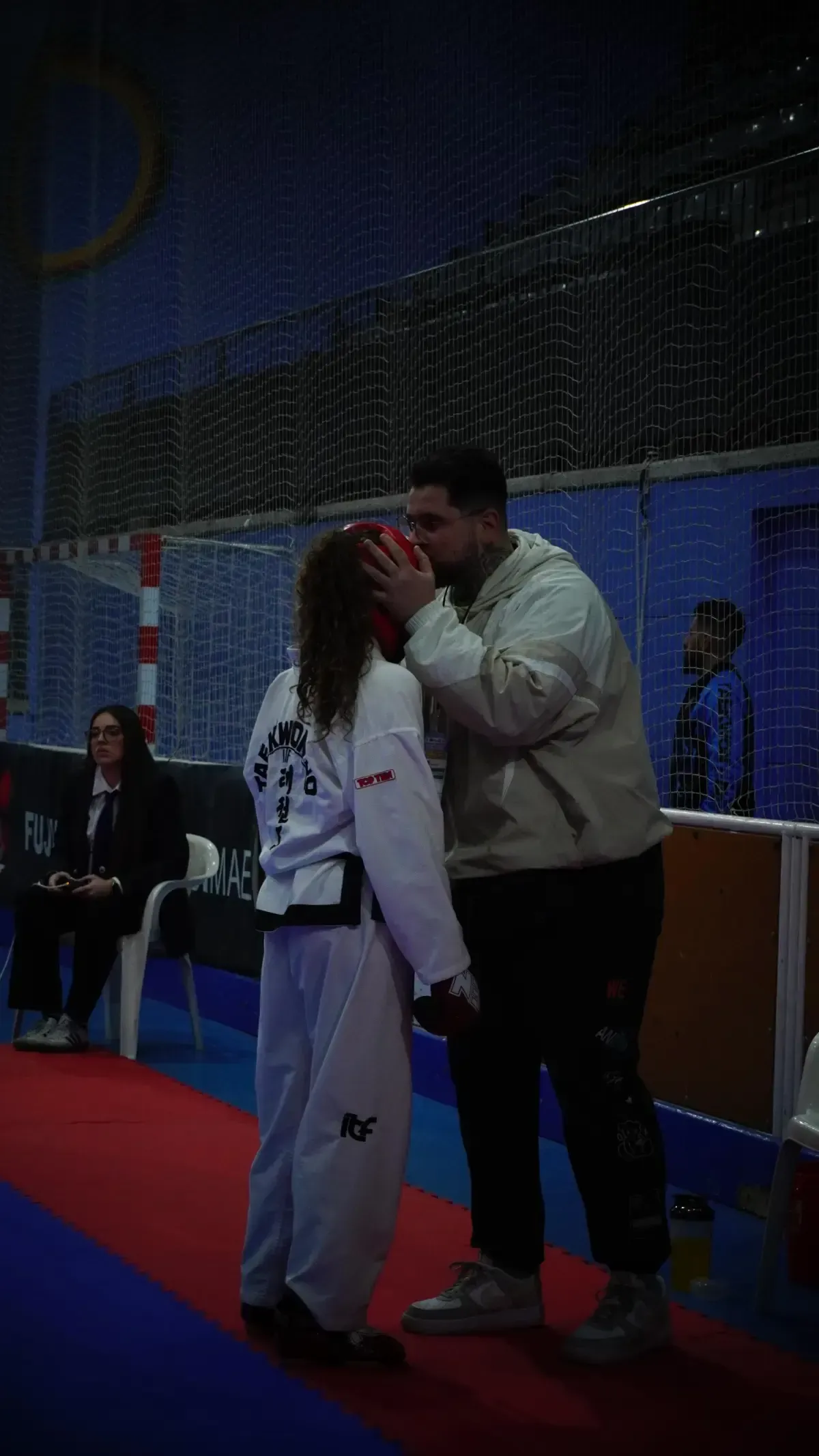 Una persona con uniforme de taekwondo permanece de pie sobre una colchoneta mientras un entrenador le besa la frente