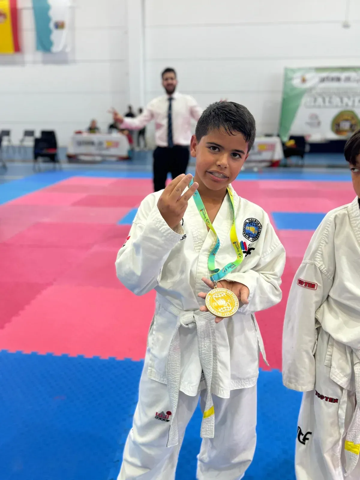 Un joven artista marcial con uniforme blanco sostiene una medalla de oro, con un instructor al fondo