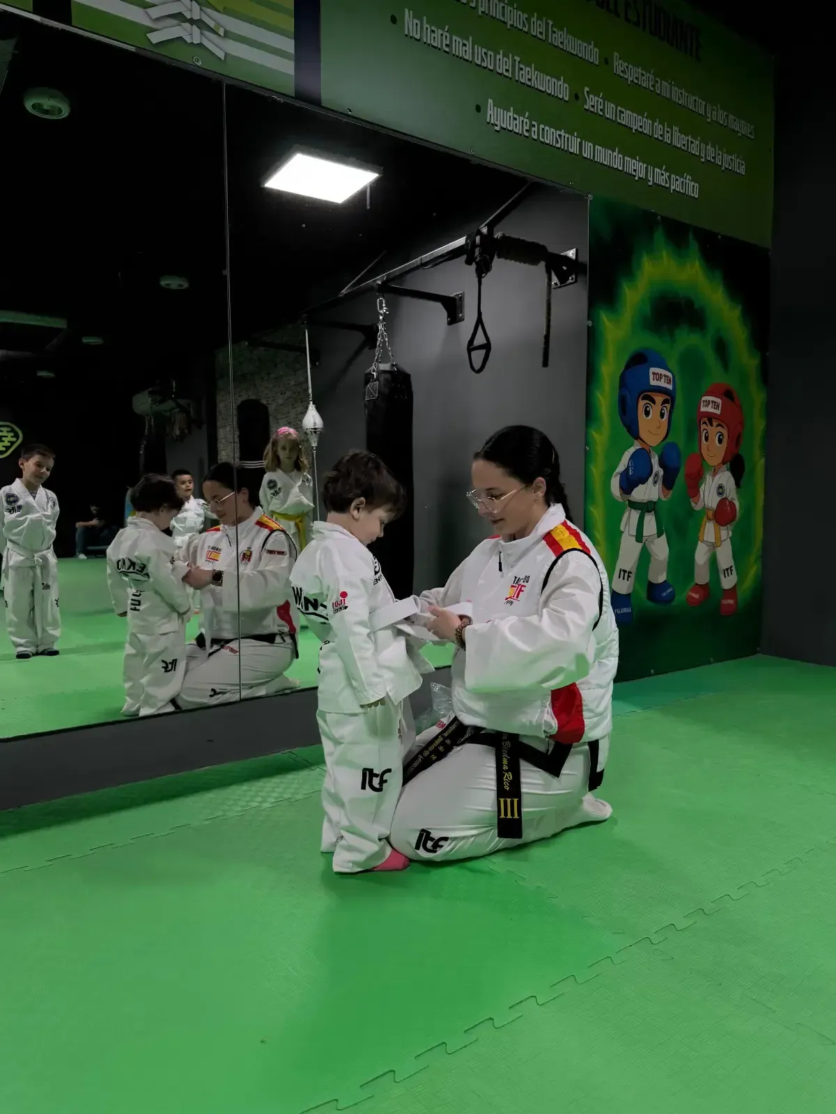Un instructor le ata el cinturón a un alumno en un estudio de artes marciales con suelo verde y murales en las paredes.