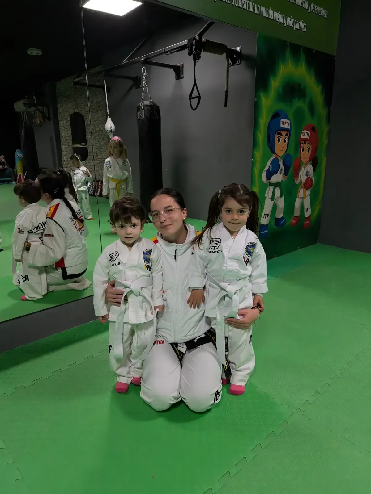 Un instructor con uniforme blanco se arrodilla entre dos jóvenes estudiantes que visten uniformes de artes marciales