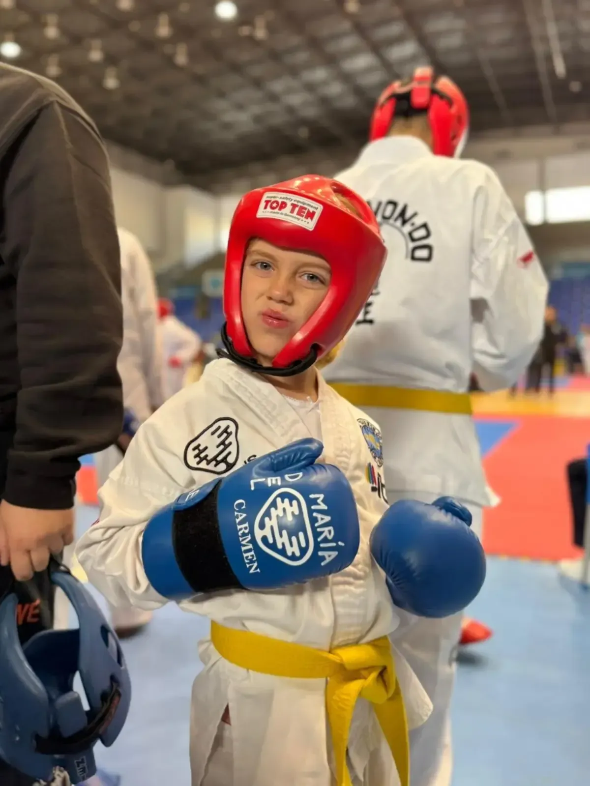 Un niño con uniforme de taekwondo, gorro rojo y guantes azules, posando con expresión concentrada en un recinto deportivo.