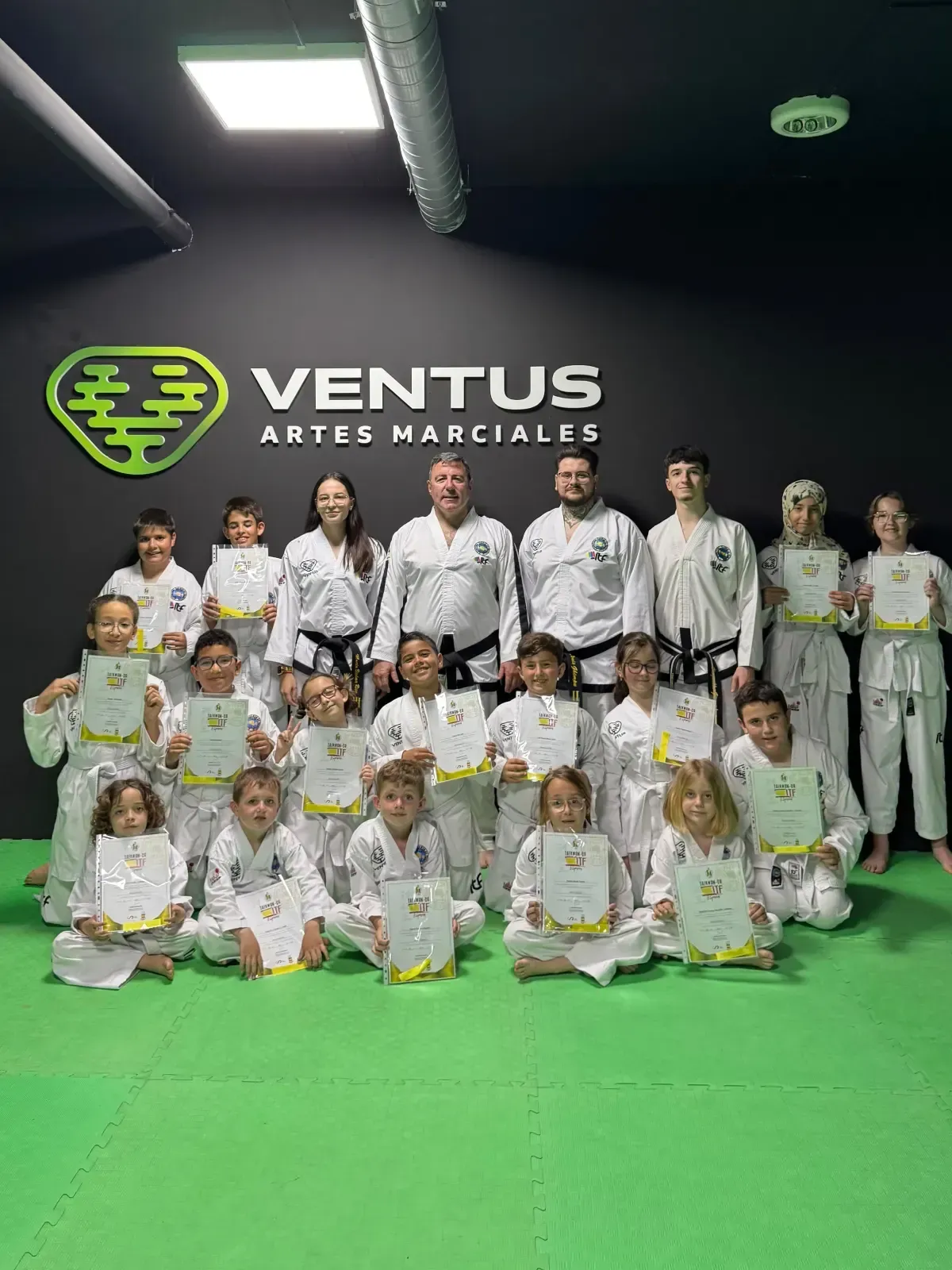 Una clase de artes marciales posa para una foto frente a un letrero de Ventus Artes Marciales; muchos sostienen certificados.