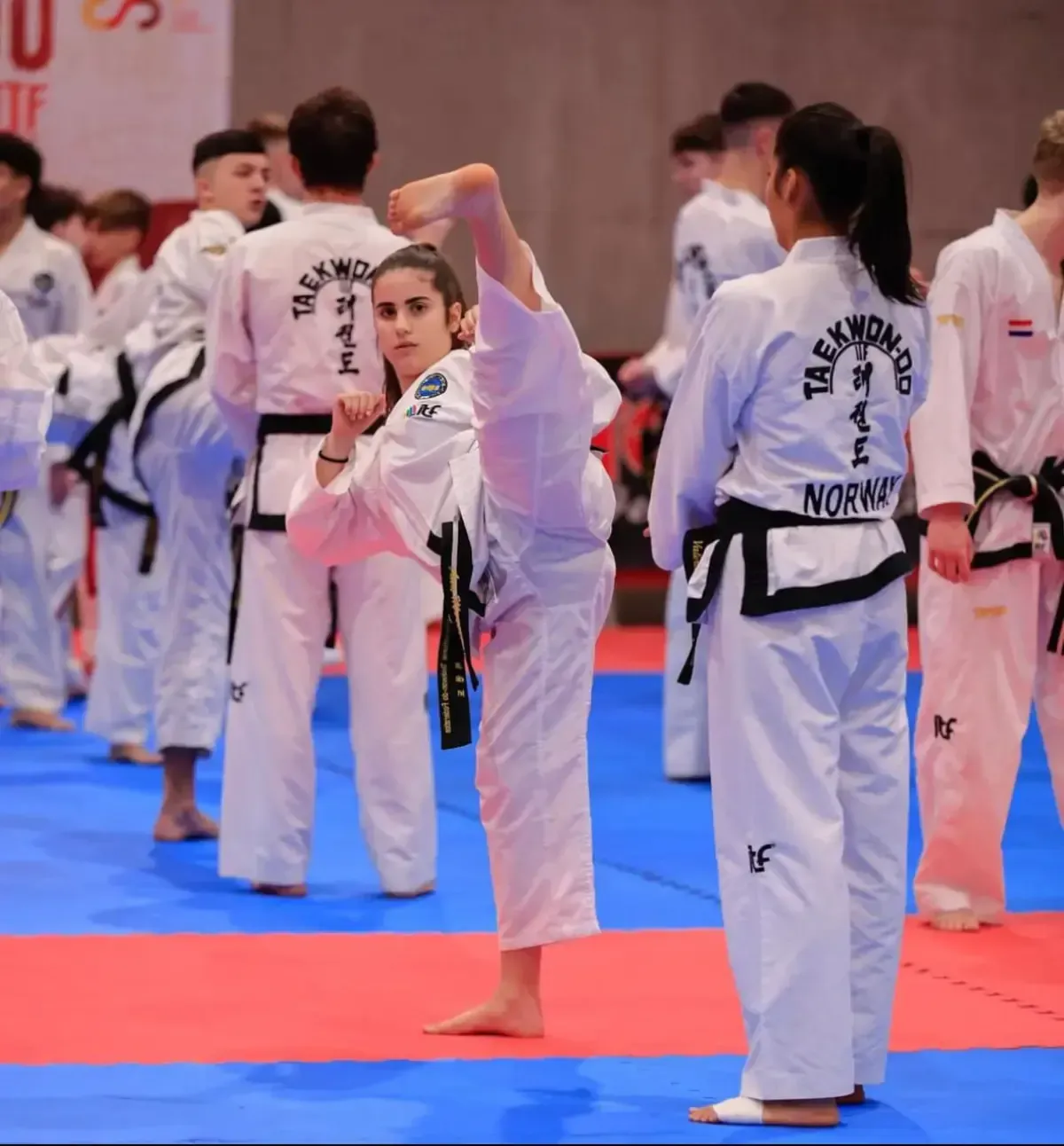Un practicante de taekwondo con uniforme blanco realiza una patada alta vertical sobre una colchoneta azul y roja
