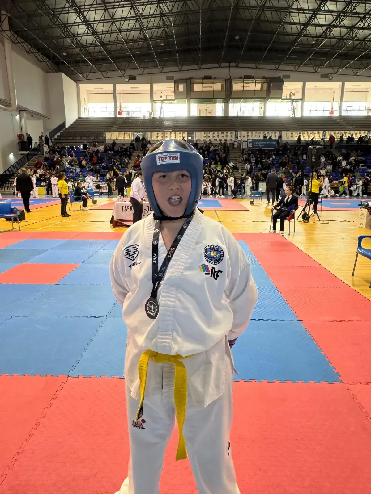 Un artista marcial con uniforme blanco, cinturón amarillo y casco azul se encuentra de pie