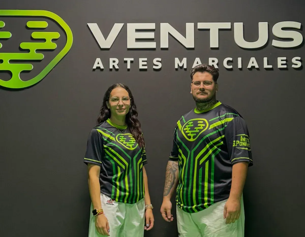 Dos personas vestidas con uniformes de artes marciales de marca posan frente a un logotipo de 