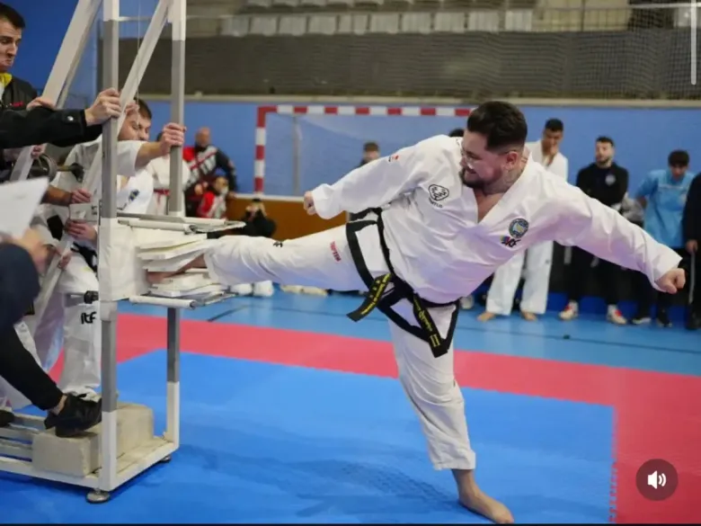 Un artista marcial con uniforme blanco realiza una patada lateral, rompiendo unas tablas de madera sujetas por una estructura