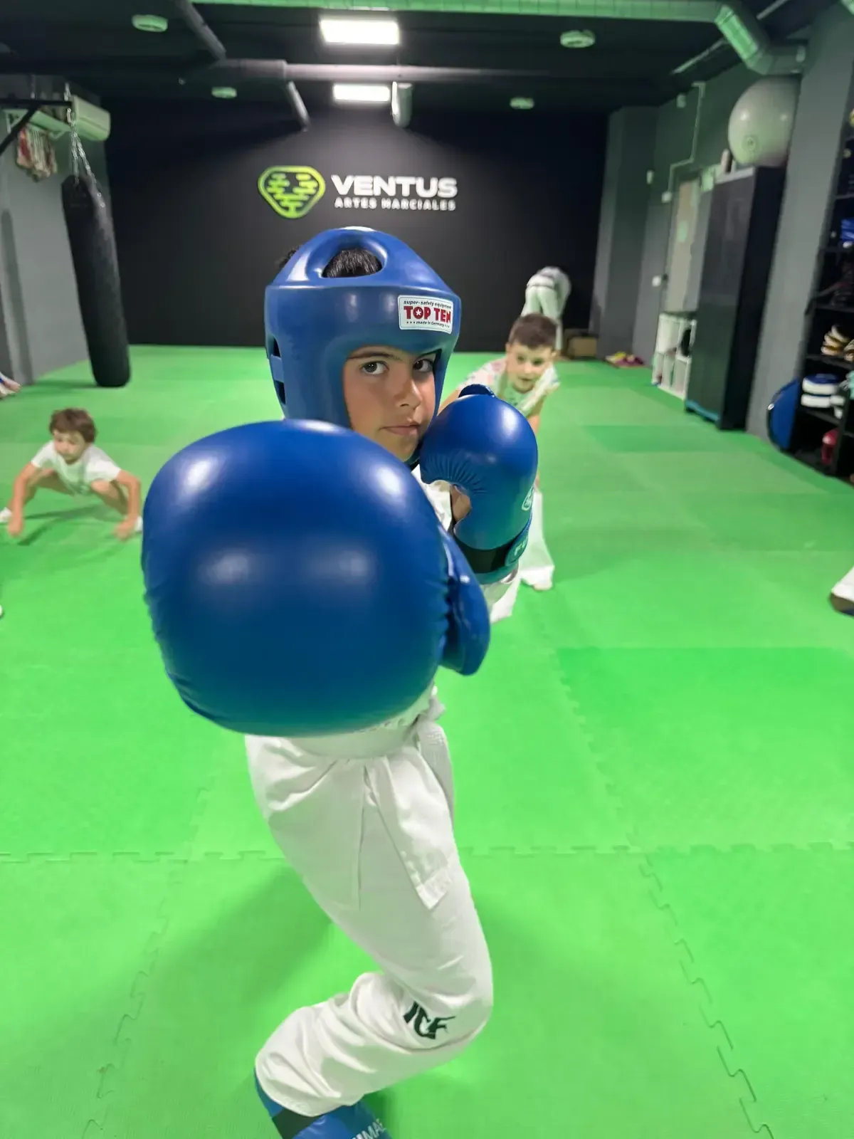 Un niño con uniforme blanco de artes marciales, casco azul y guantes de boxeo posa en un gimnasio