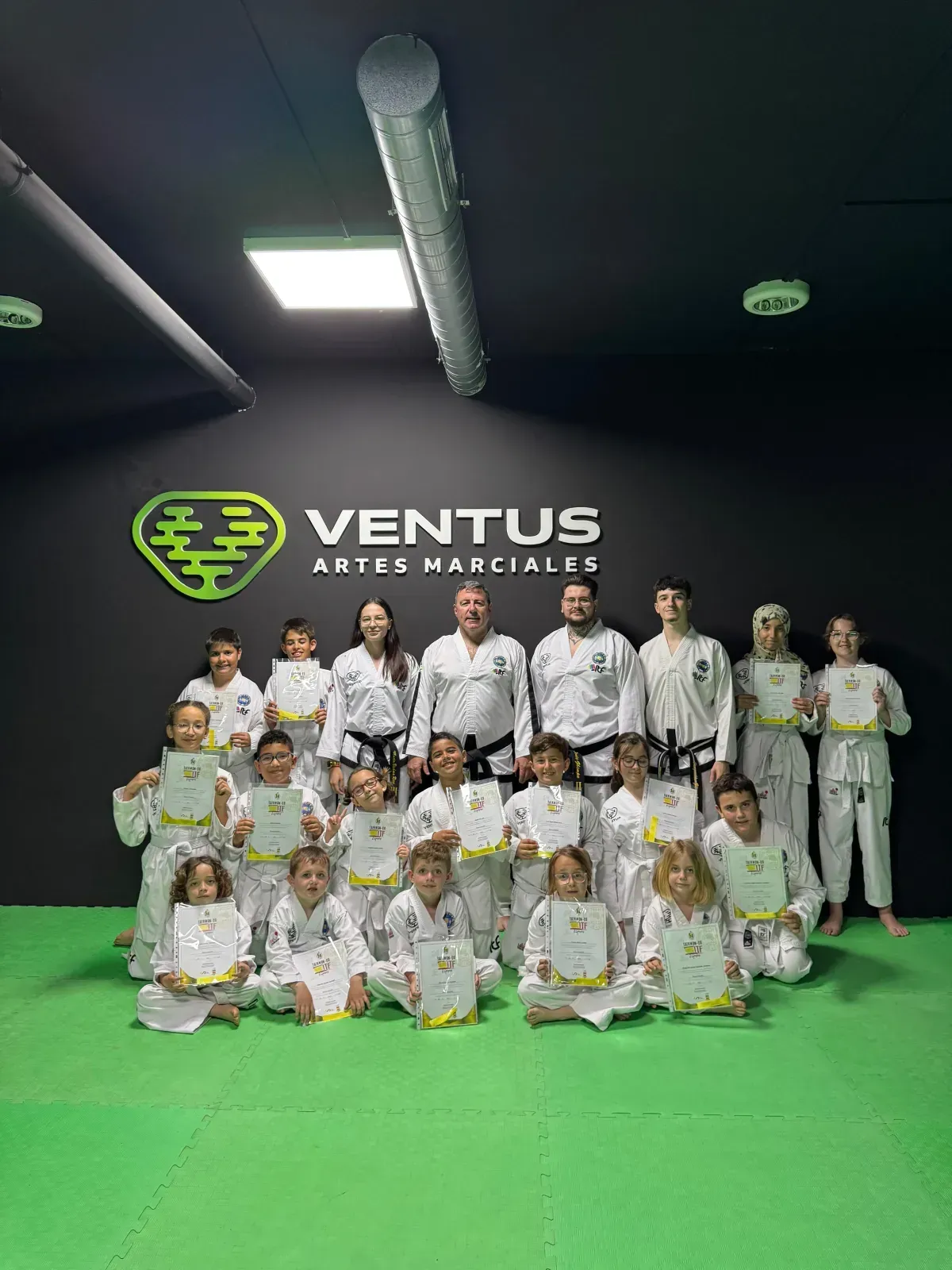 Un grupo de artistas marciales con uniformes blancos sostienen certificados frente al logotipo del gimnasio